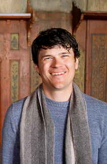 Man in light blue sweater, brown scarf, wood panel in background